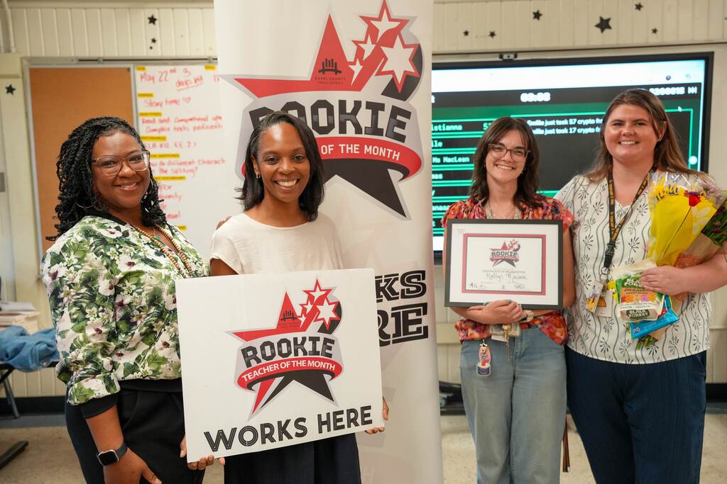 Four women stand in a room. One holds a certificate, another holds flowers. The third holds a sign reading "Rookie Works Here." A backdrop displays "Rookie of the Week."