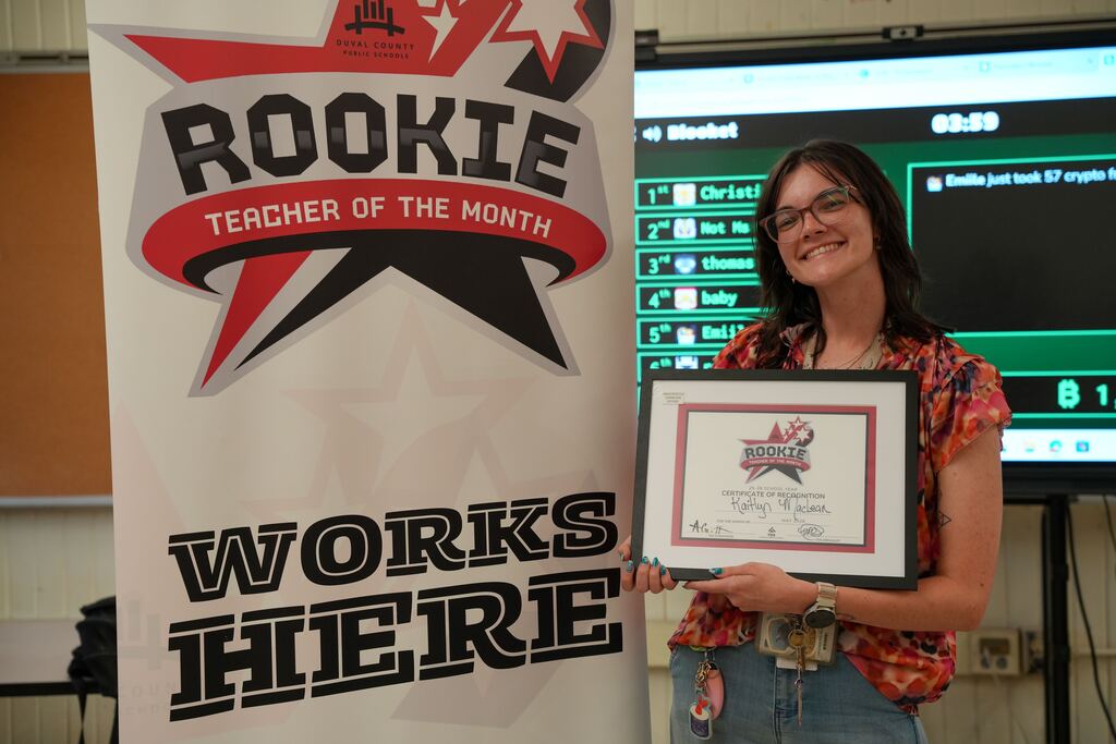 Woman stands next to a banner and holds a certificate, smiling. A monitor with a game displays behind her.