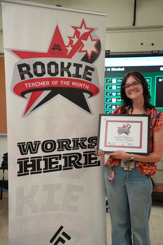 Person holding a framed certificate with a "Rookie Teacher of the Month" banner behind them.
