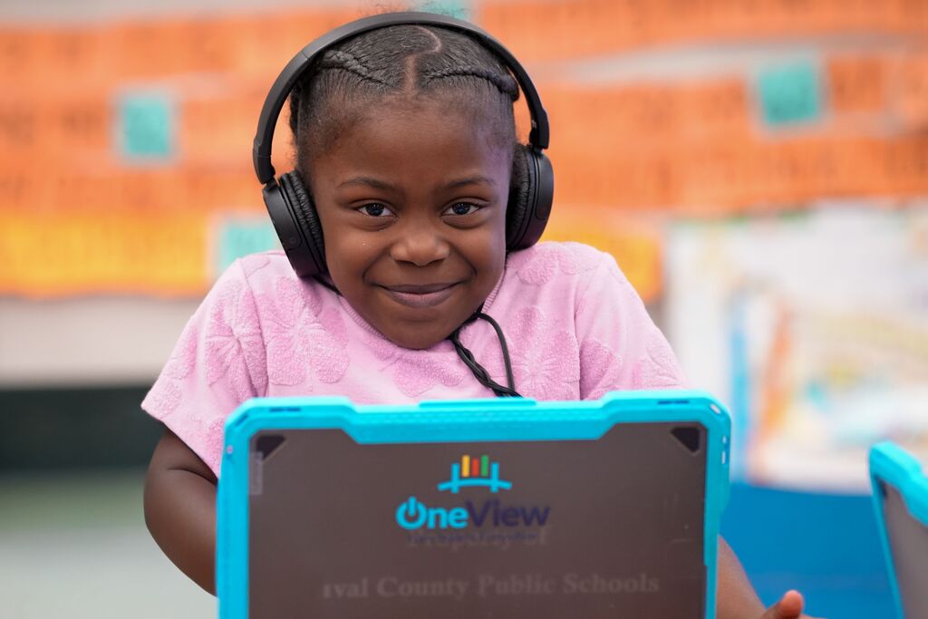 A young girl in pink headphones smiles, holding a tablet displaying "One New" on a white background.