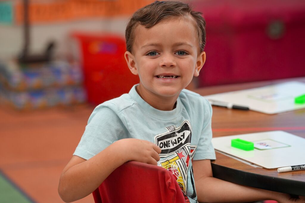 A young boy sits in a chair at a desk, wearing a light blue T-shirt with a graphic design.