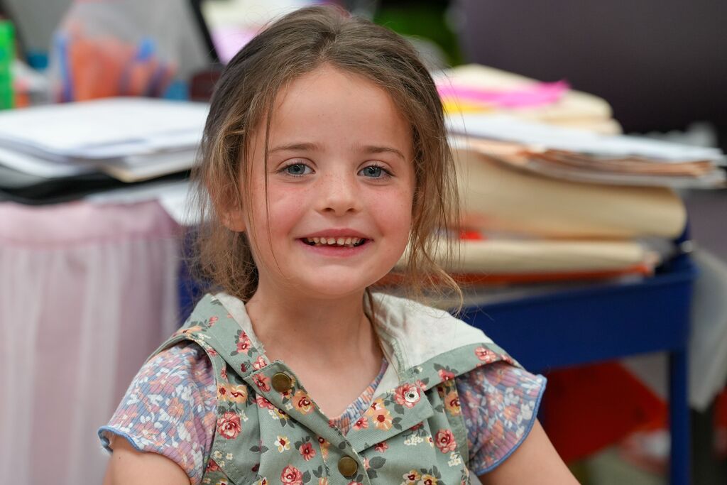 Young girl wearing a floral dress smiles, with a table and papers in the background.