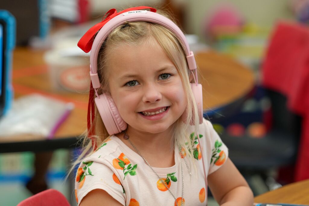 A young girl in pink headphones smiles in a classroom with a desk and chairs behind her.