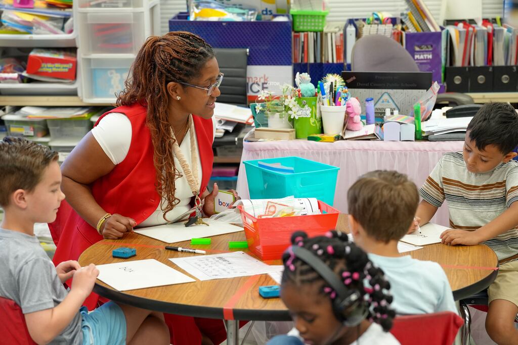 A teacher in red assists children seated at a round table in a classroom with papers, pencils, and other items.