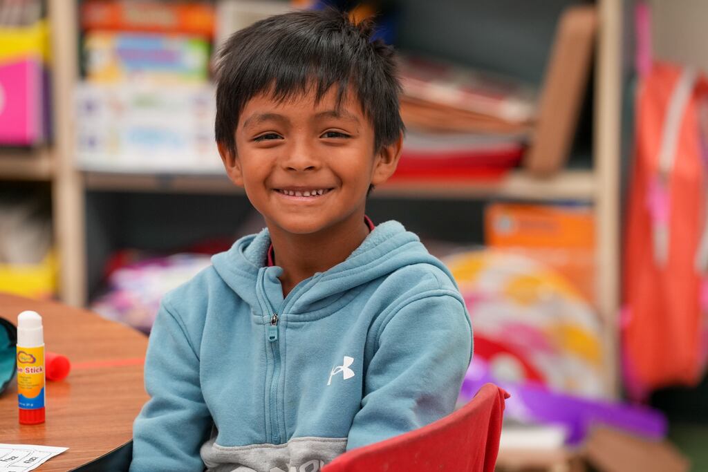A young boy with a broad smile, wearing a blue hoodie, sits at a red chair in a classroom.