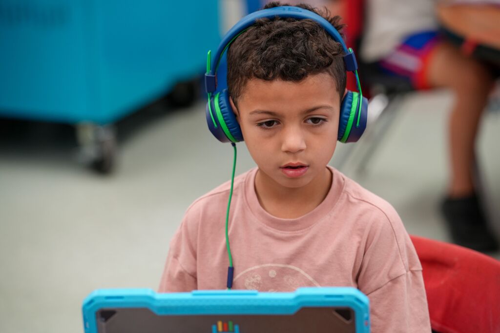 Child wearing headphones, using a tablet with a colorful display, sitting on a red chair.