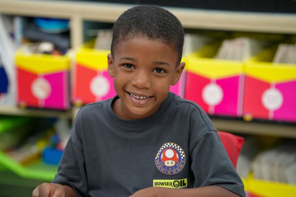 A young child with short hair, wearing a gray shirt with a logo, sits in a classroom.