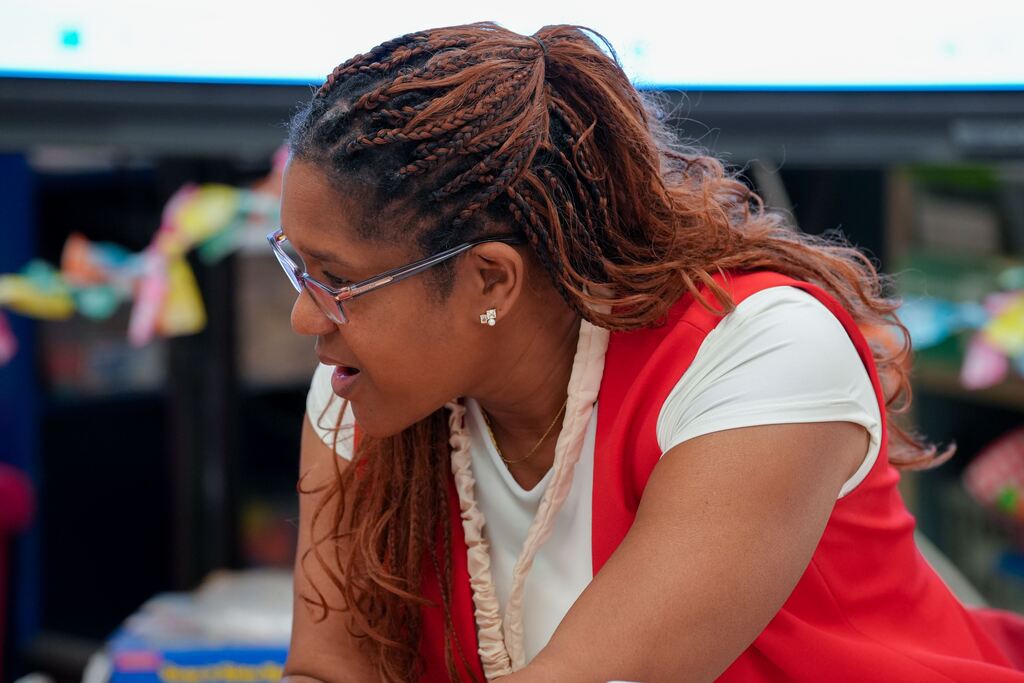 A woman with long hair, wearing glasses, and a red vest looks downward in a classroom.