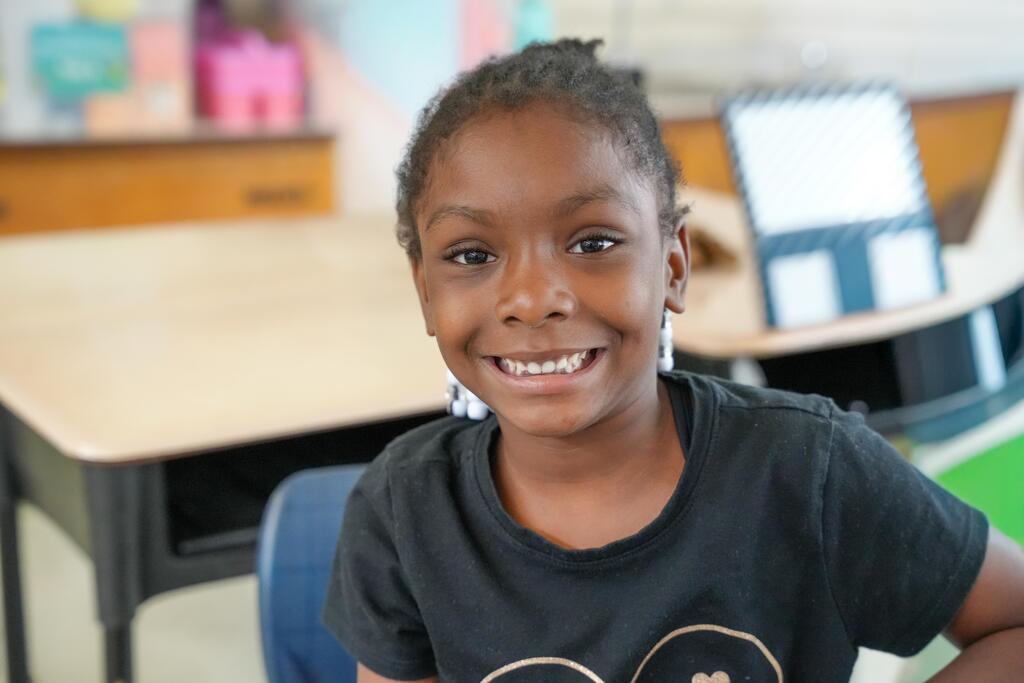 A smiling child in a black t-shirt sits in a classroom. Desks and a pink wall are in the background.