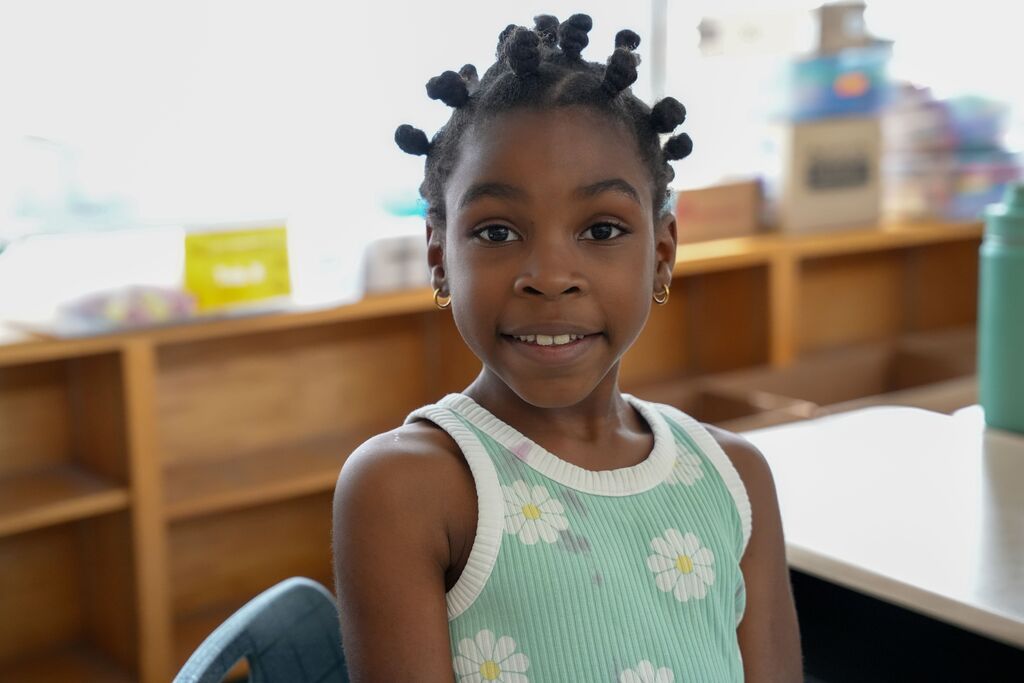 A smiling girl with pigtails sits in a chair in front of shelves and a desk.