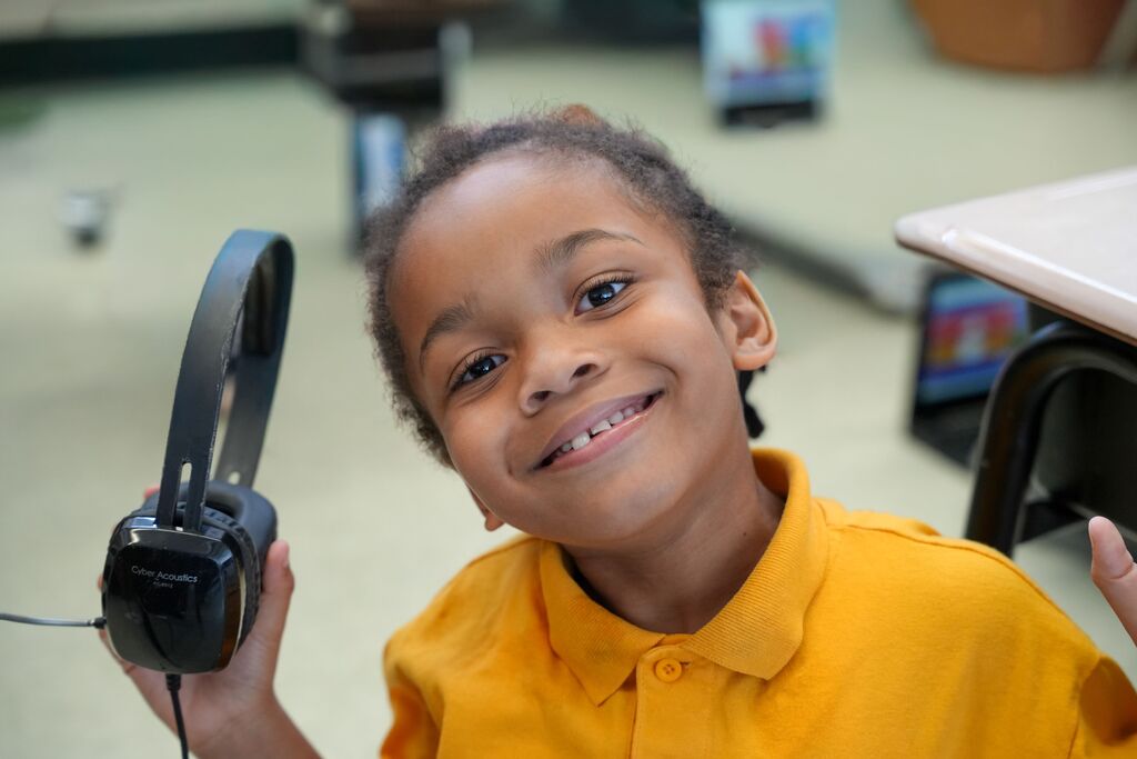A child wearing headphones holds them up while smiling, seated in a classroom with a table in the background.