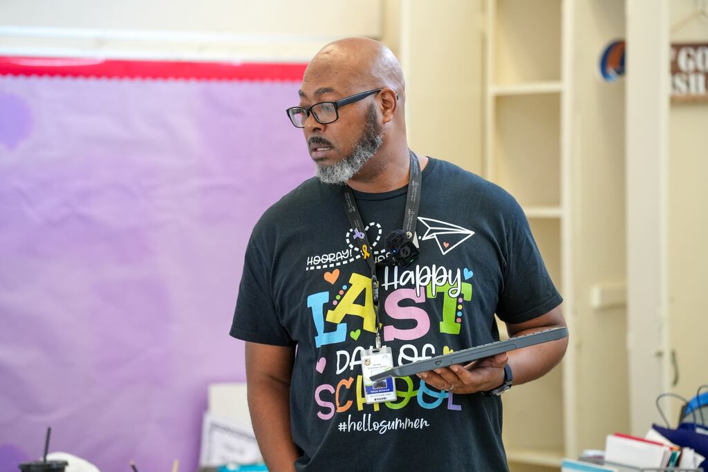 Man in glasses, wearing a black T-shirt, holding a tablet, standing in front of a classroom.