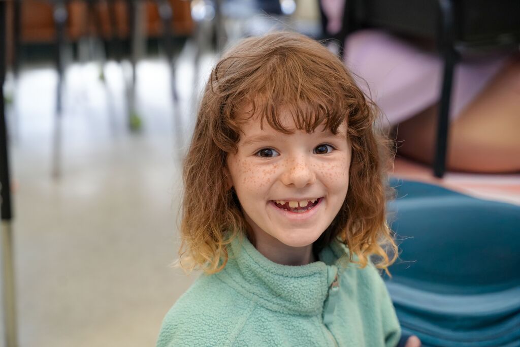 A child with curly brown hair, wearing a green shirt, smiling and looking at the camera in a classroom.