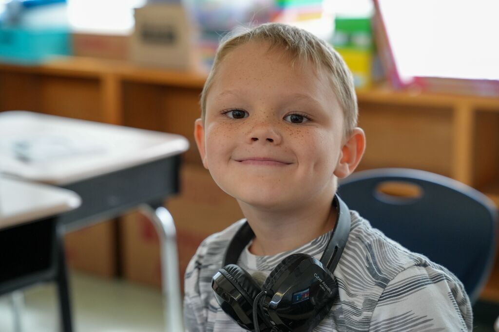 A smiling boy wearing headphones, seated in a chair in a classroom with a desk behind him.