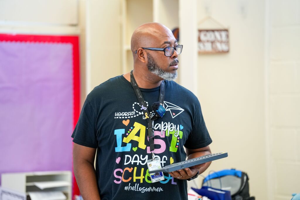 Man in glasses stands in classroom, holding tablet, wearing a black shirt with colorful text, standing before white wall.