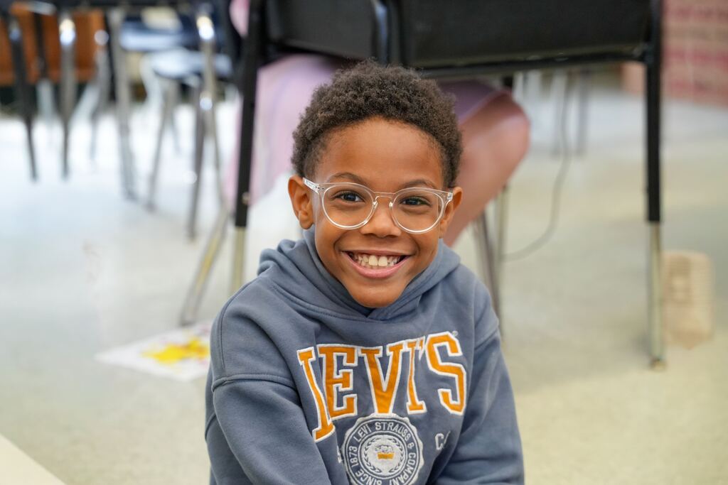A young boy with short hair and glasses smiles in a classroom, wearing a gray hoodie with "LEVIS" printed on it.