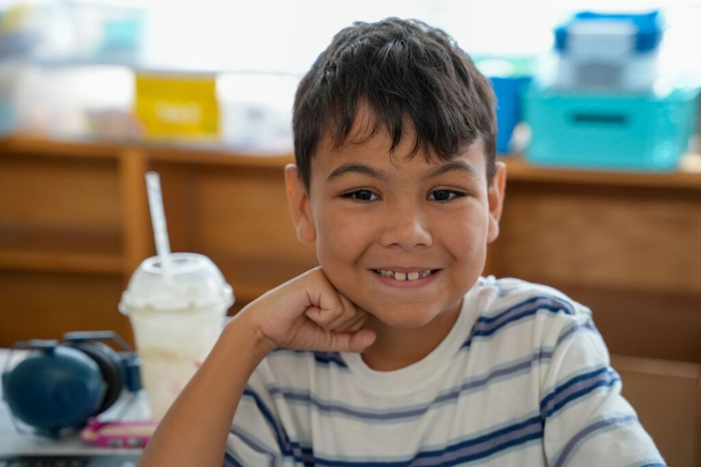 Boy in a classroom, smiling with his chin on his fist, seated at a desk with headphones and a beverage.