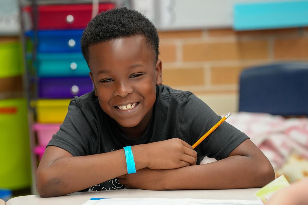 A smiling boy sits at a table in a classroom, holding a pencil, with colorful storage boxes and brick walls in the background.