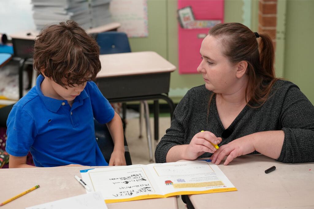 A young boy and a woman sit at a desk. The boy wears a blue shirt; the woman wears a black shirt. They study a book.