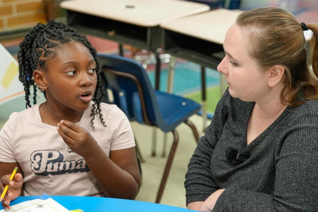 A young girl in a pink shirt looks at a woman in gray, seated at a table.