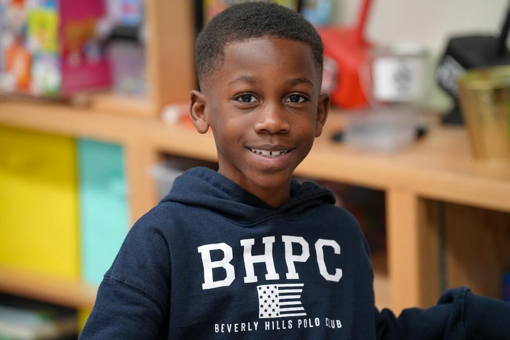 A smiling young boy in a navy hoodie with the text "BHPC" and a flag on it. He is in a classroom with shelves behind him.