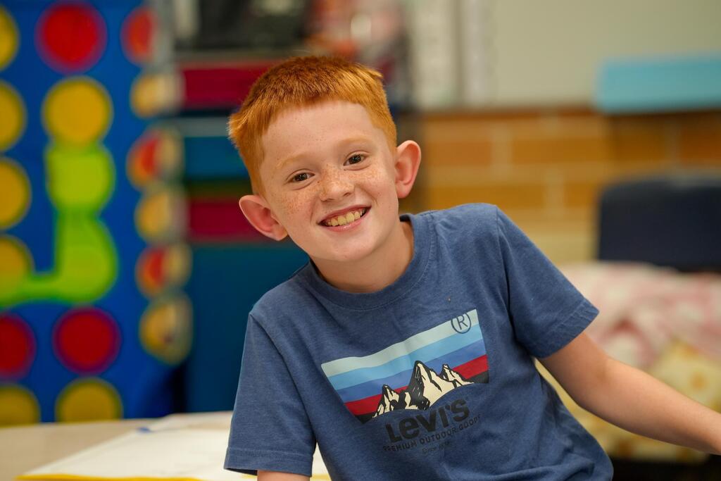 A boy with red hair smiles in a colorful room with a brick wall, wearing a blue shirt with the word "Levi's".