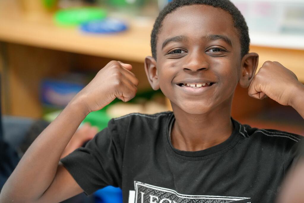 A young boy poses with his hands on his head and biceps flexed, smiling brightly in a classroom.