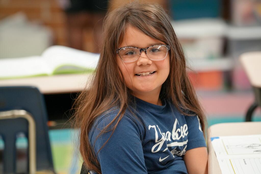 A smiling child with glasses and a blue shirt sits at a desk in a classroom.