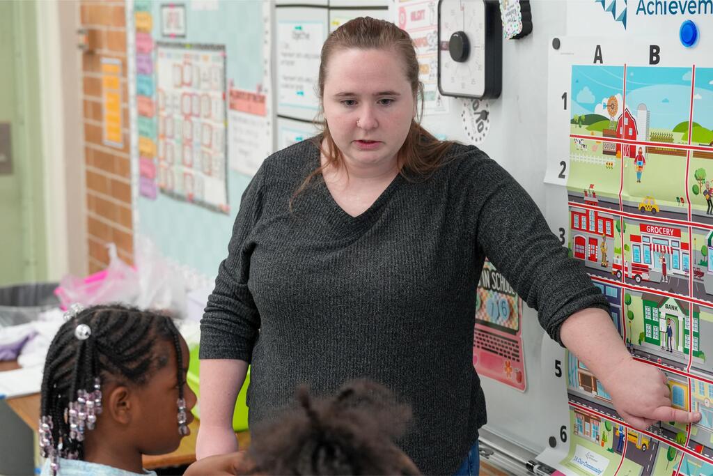 Woman in gray shirt standing, speaking to a girl with braids. Background includes a bulletin board and educational materials.