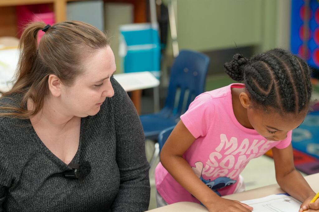 An adult and a child sit at a desk. The adult watches the child work on a paper.
