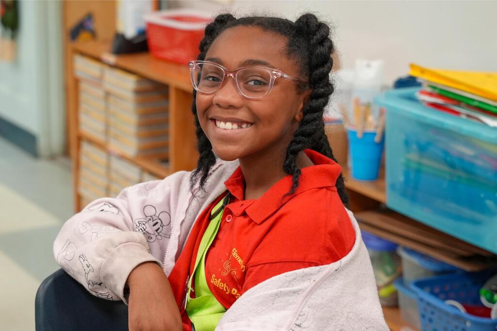 A girl in a red shirt and glasses sits in a classroom with shelves and storage boxes in the background.