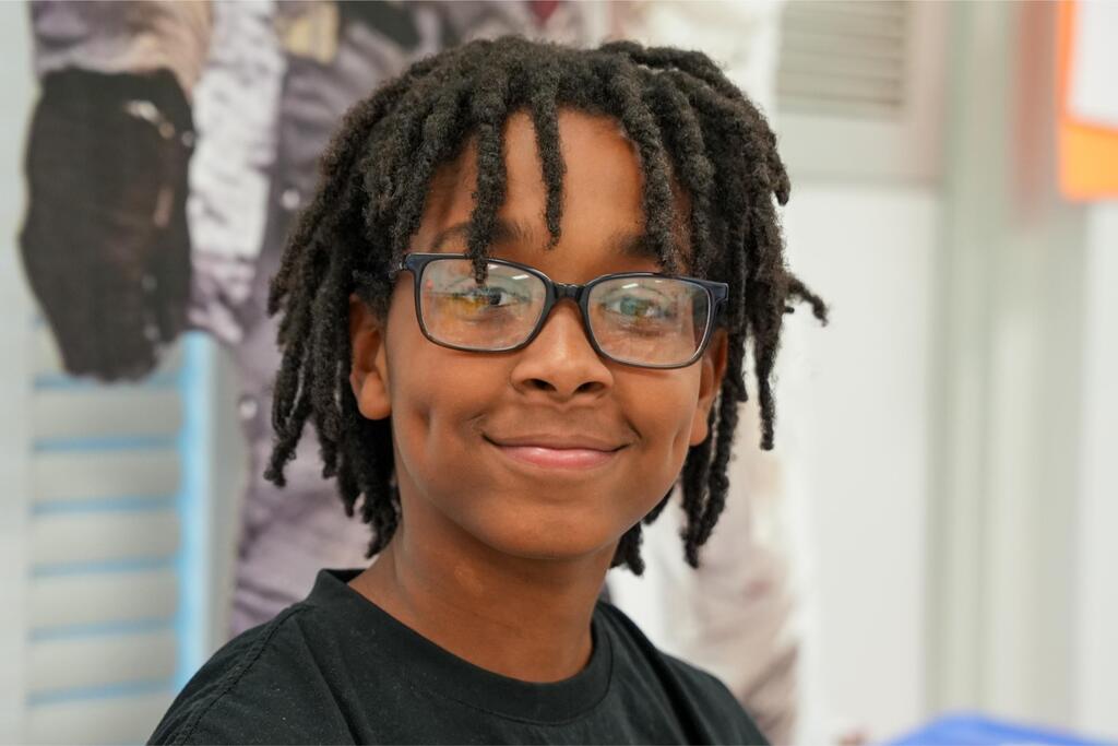 A person with short, braided hair and glasses smiles indoors. They wear a black T-shirt.