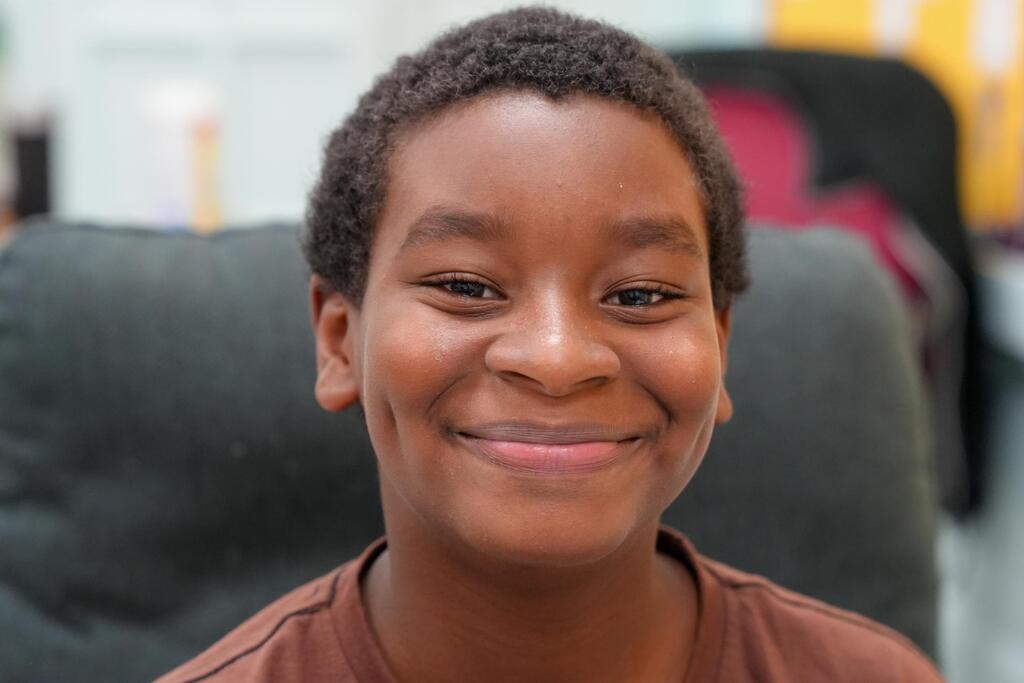 A smiling child with short hair, wearing a brown shirt, sits in a chair. Blurry background includes a chair and other objects.