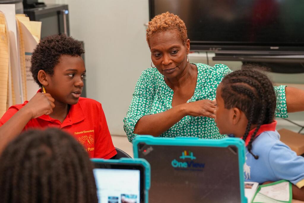 A woman in green stands by a table, teaching two children seated. They are in a classroom with laptops.