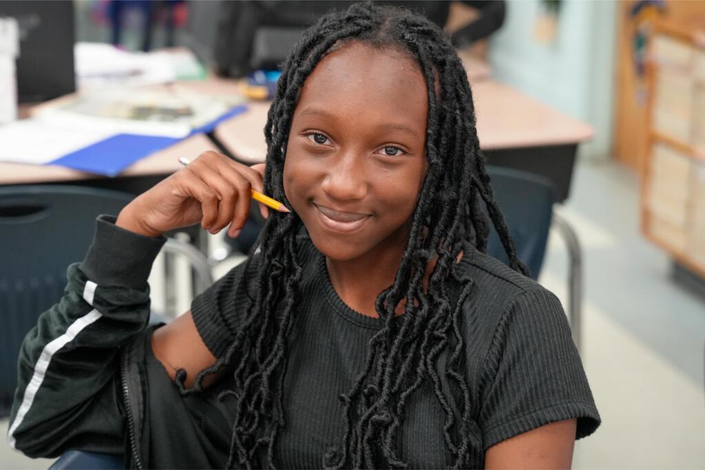 A woman with braided hair holding a pencil and smiling, seated in a classroom with a desk behind.