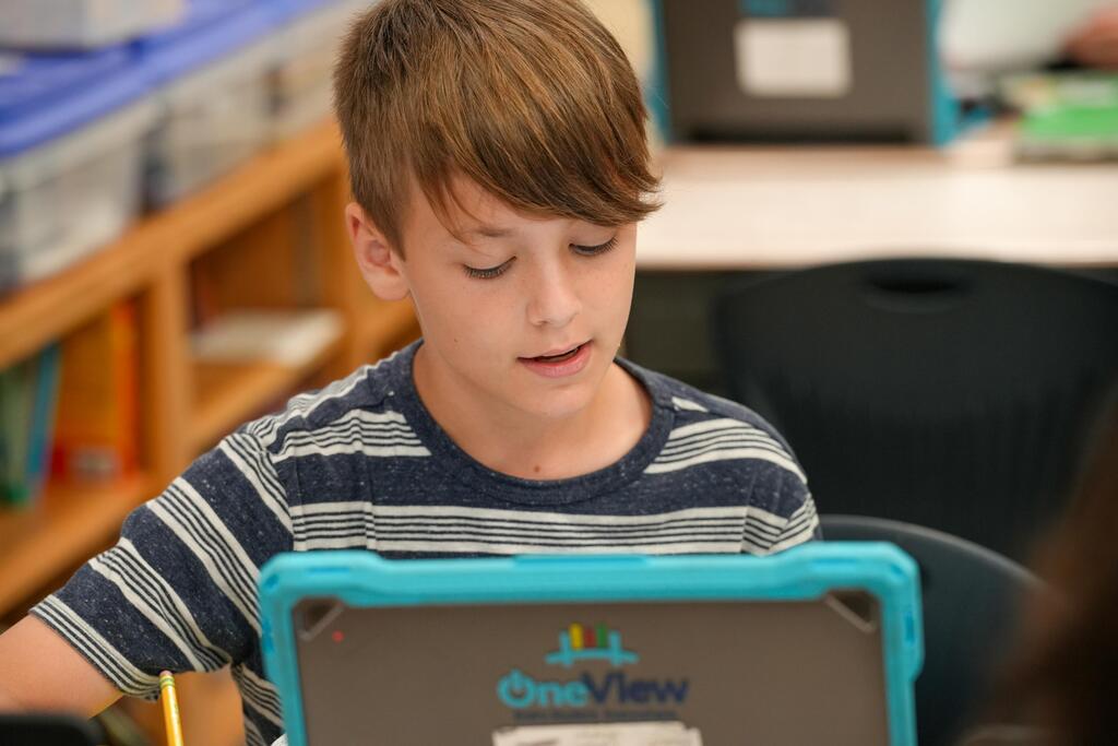 A boy with short hair wearing a striped shirt uses a tablet in a classroom.