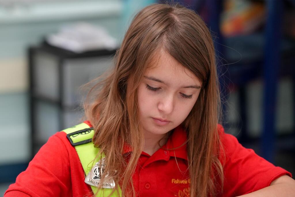 A girl with long hair wearing a red shirt and a yellow safety vest sits at a desk.