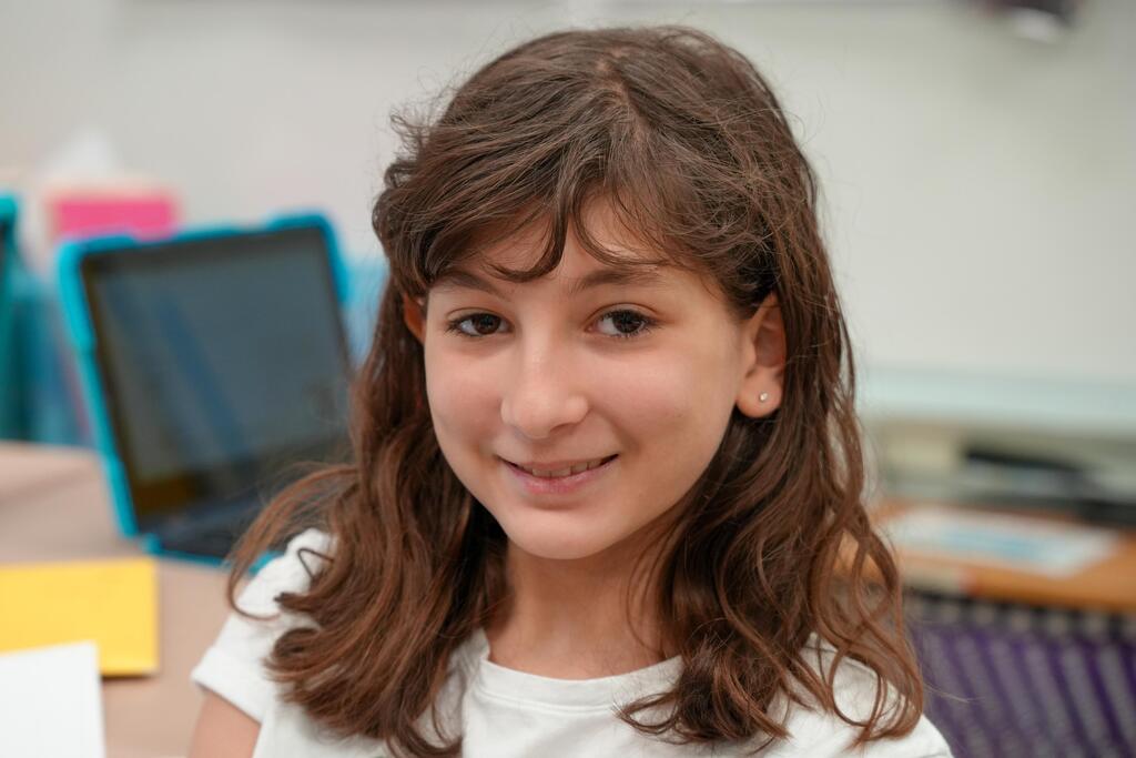 A girl with brown wavy hair smiles in a classroom. She wears a white shirt and sits near a computer and various items.