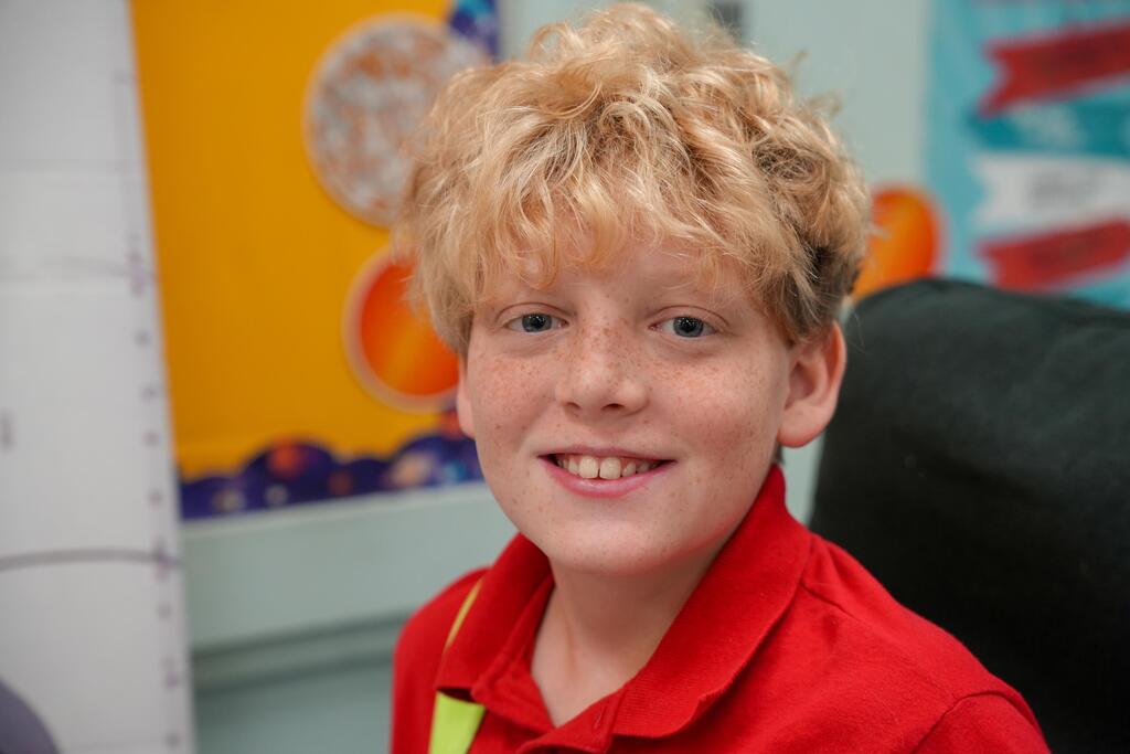 A boy with short blonde hair in a red shirt smiles in a classroom. Behind him are posters with colorful shapes.