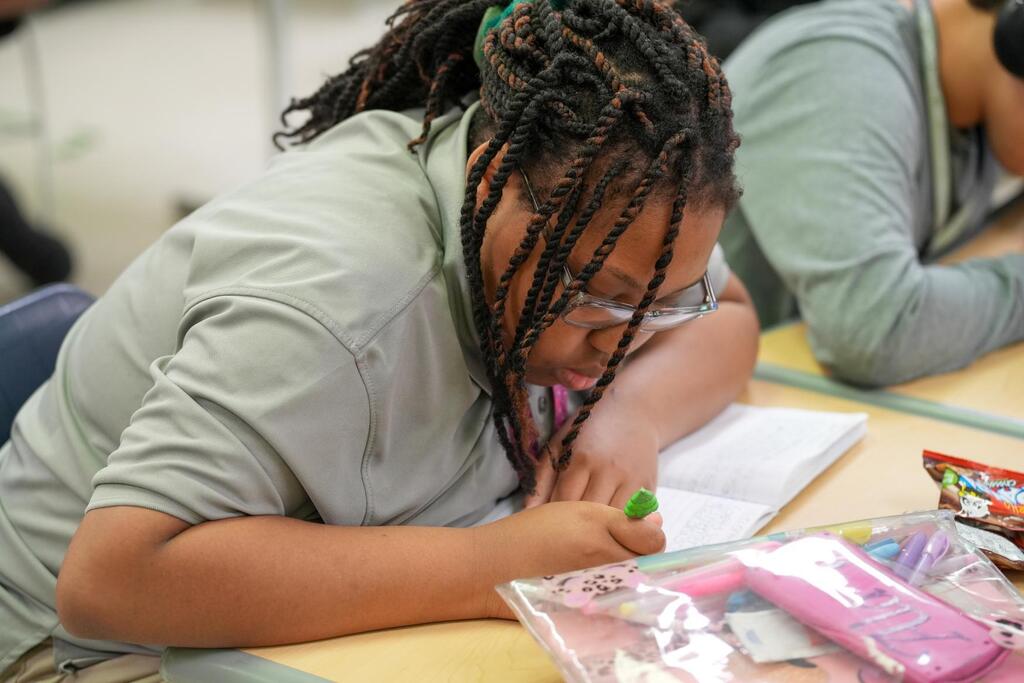 A student focuses on writing in a notebook while seated at a desk, with a backpack and snacks nearby.