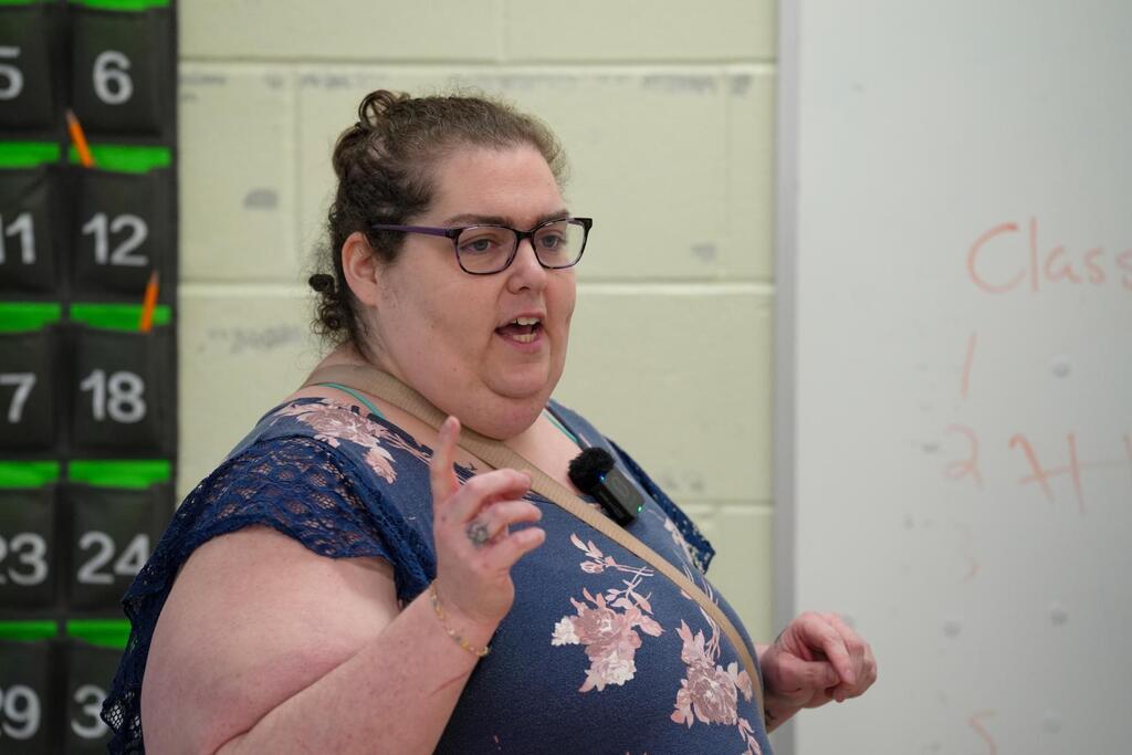A person speaks into a microphone. A whiteboard behind them has writing. A calendar is to the left.