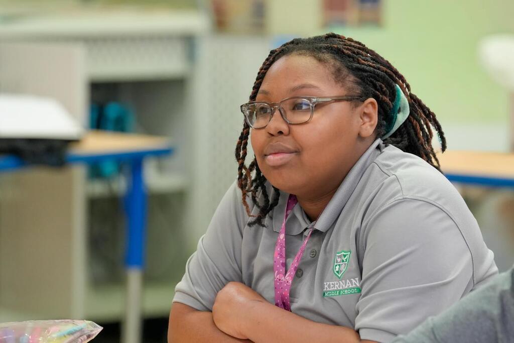 A person with glasses and a gray shirt sits in a classroom. A blue desk is behind them.