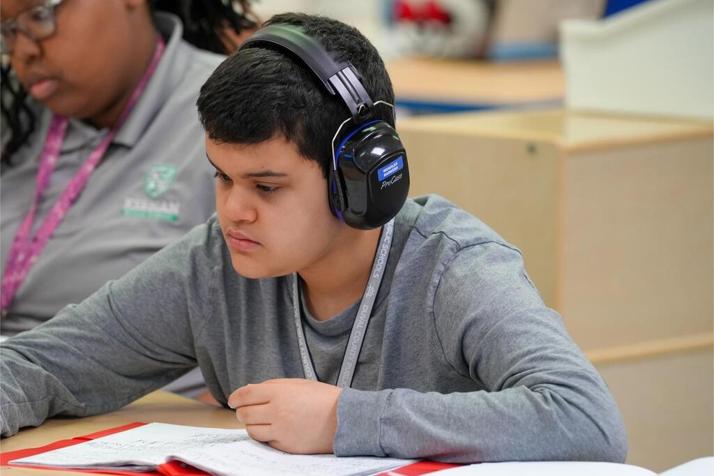 A young man wearing headphones sits at a table, studying. Another person sits behind him.