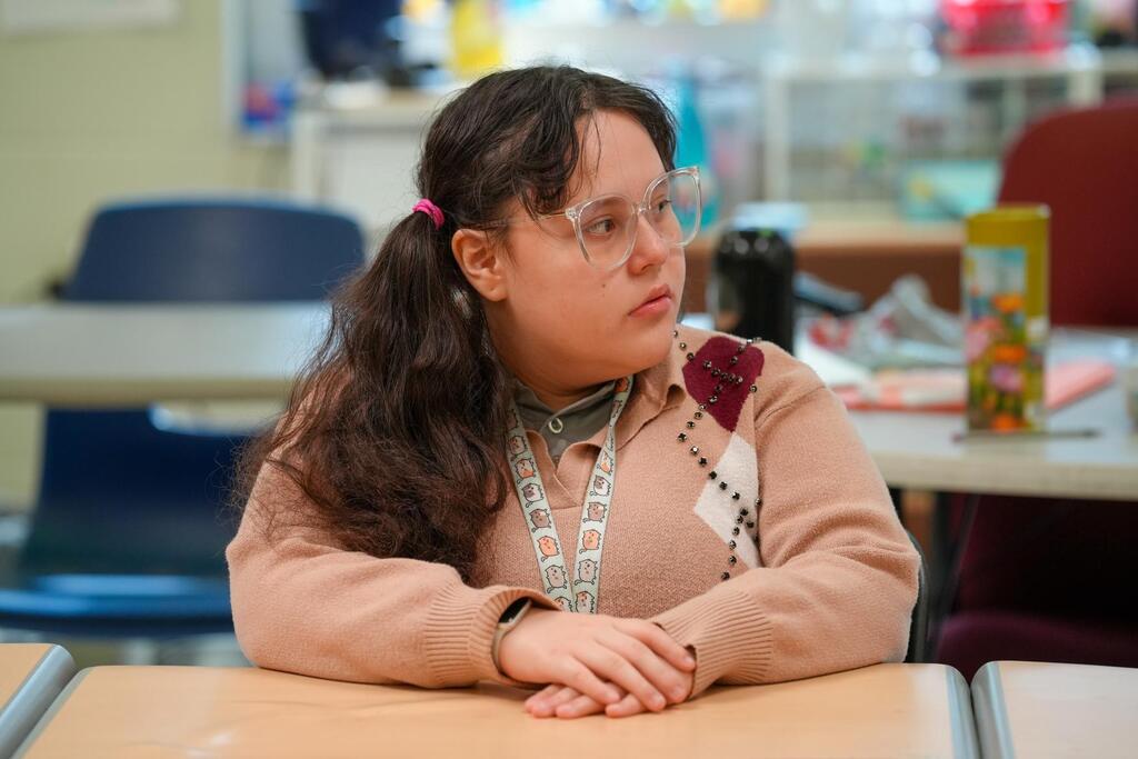 A person with glasses and a ponytail sits at a desk in a classroom with their arms folded.