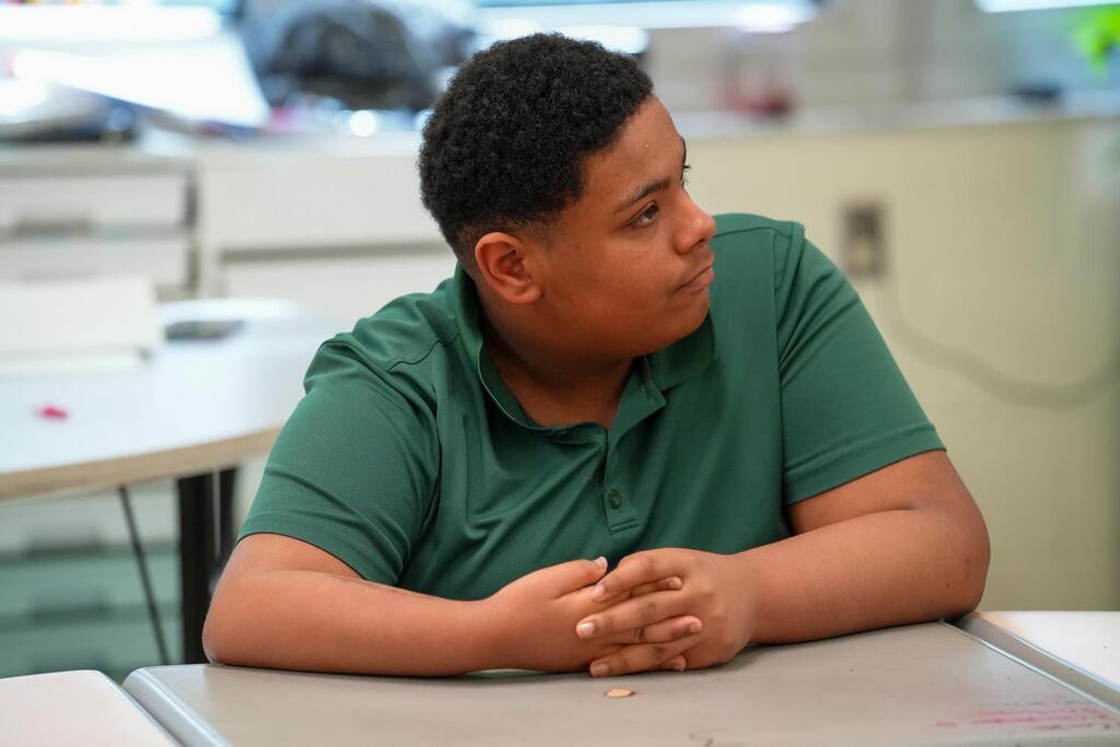 A young person in a green shirt sits at a table, looking downward, with their arms crossed.