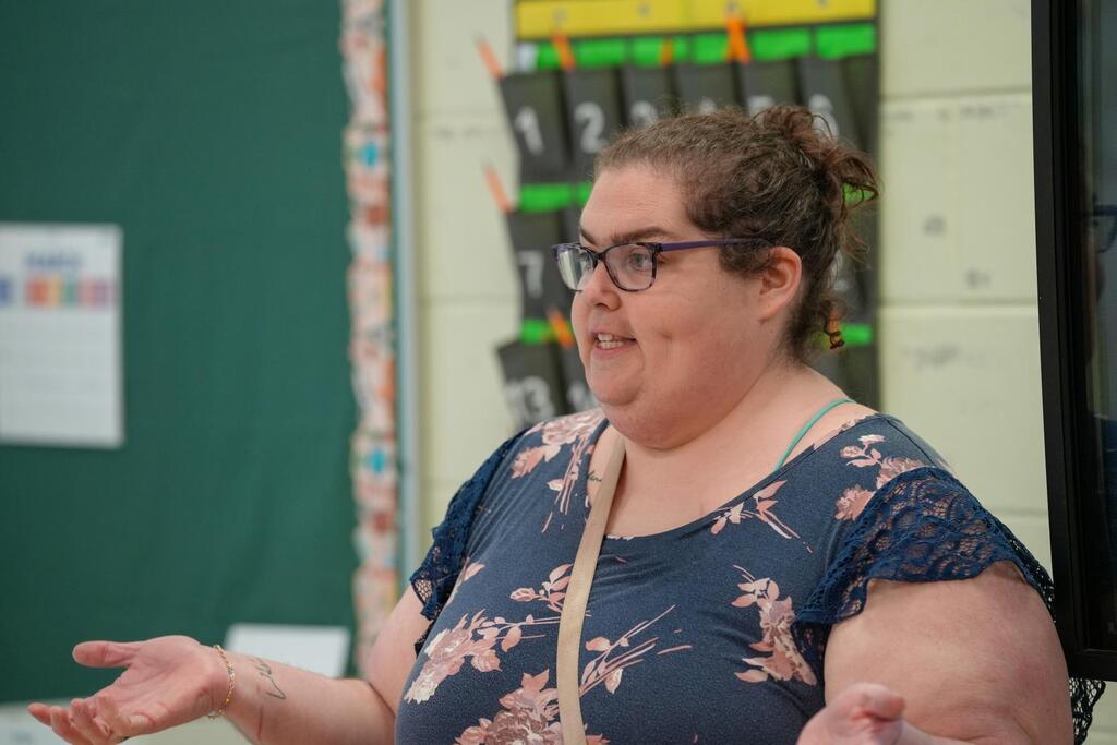 A woman in glasses and a floral shirt stands in front of a classroom with a green wall and numbered folders.