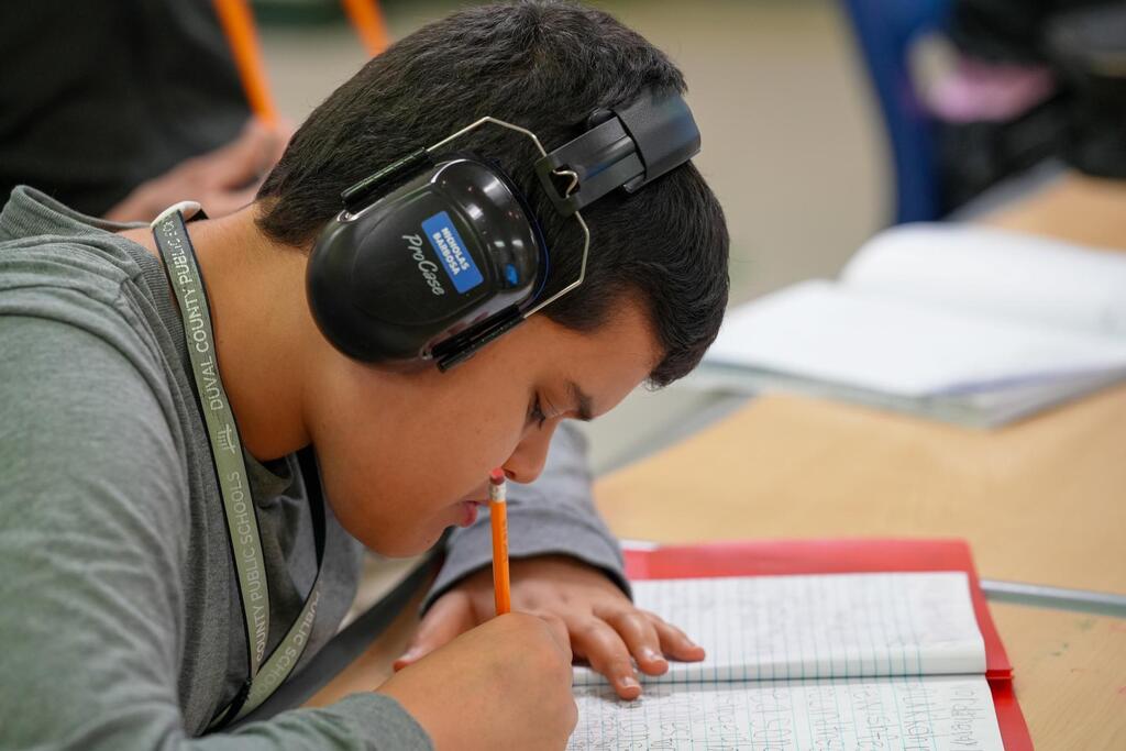 A student with headphones on, writing in a notebook at a desk with papers and pencils.