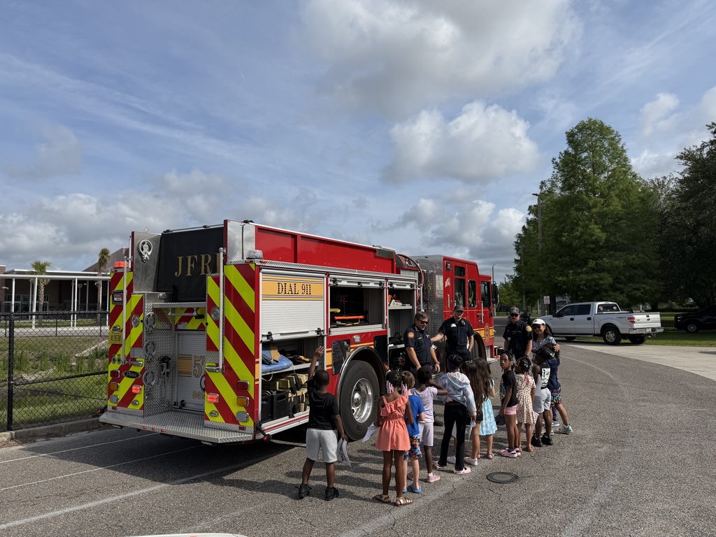 A group of students stands in front of a fire truck, listening to an information session from a group of firefighters. 