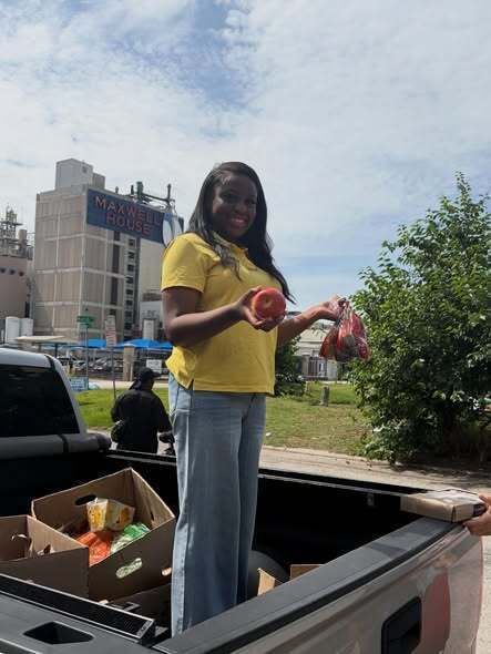 A student stands smiling in the back of a truck, holding a bag of apples and surrounded by other fresh produce as part of a food drive.