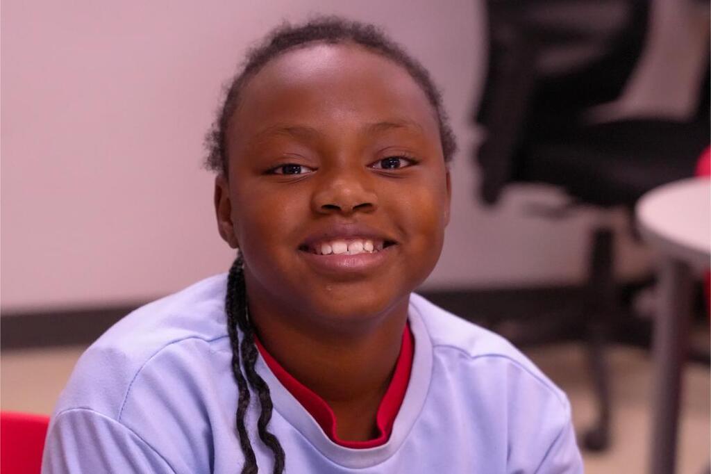 Person with braided hair in white and red shirt sitting in a chair and smiling.